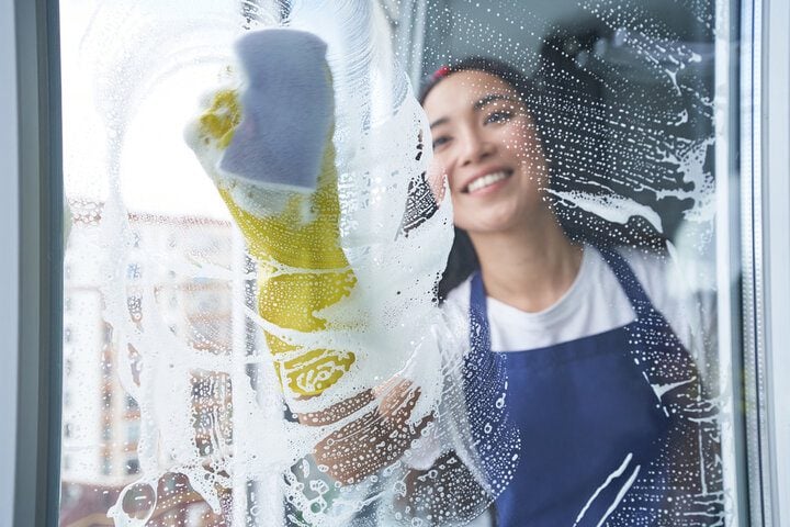 Smiling woman cleaning a glass window with a soapy sponge, wearing a blue apron and yellow gloves, with a background view of buildings blurred through the glass.